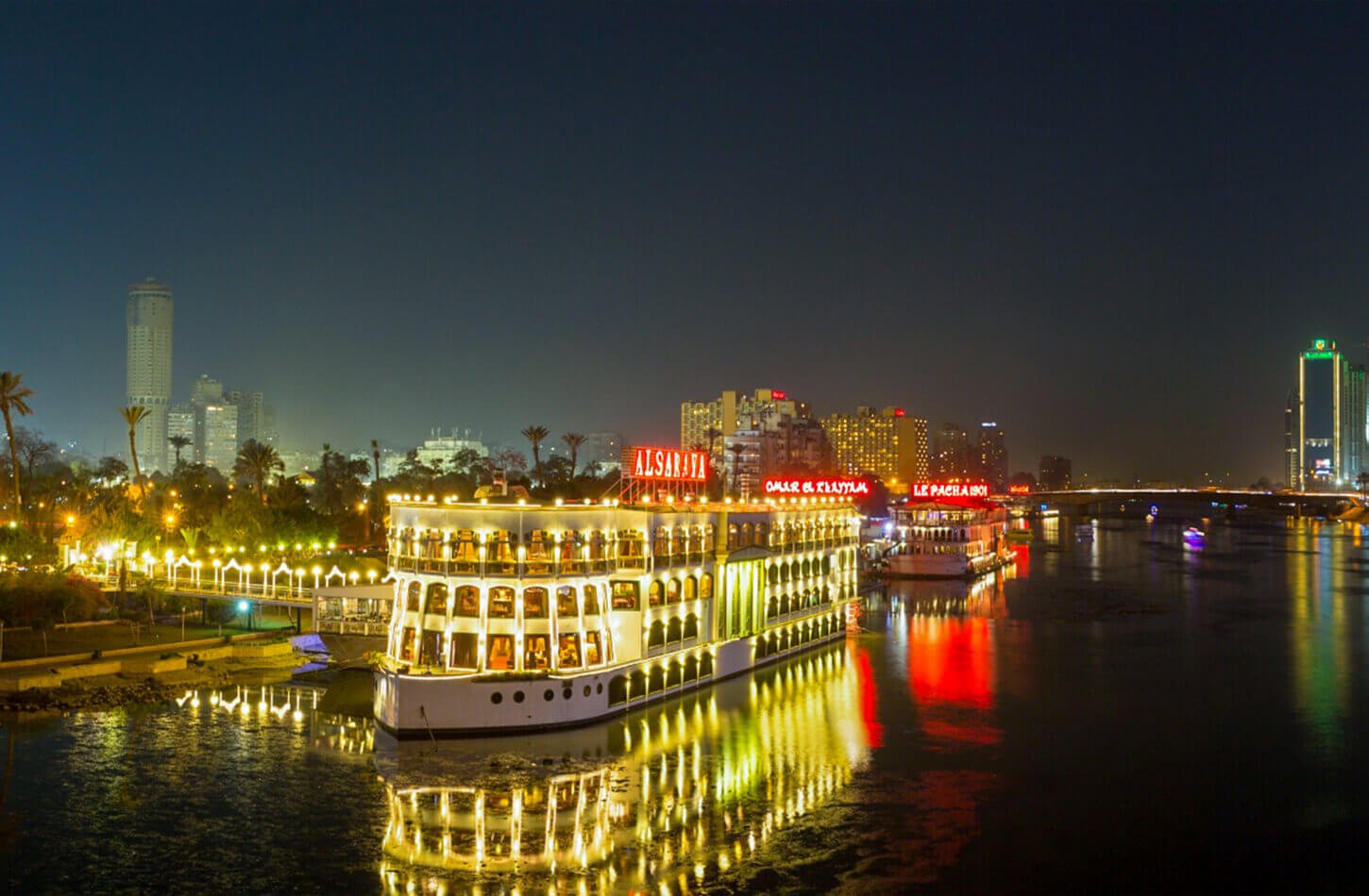 A brightly lit cruise boat docked along the Nile River in Cairo at night, reflecting vibrant gold and red lights onto the water's surface, highlighted as one of the best Things to Do in Cairo at Night.