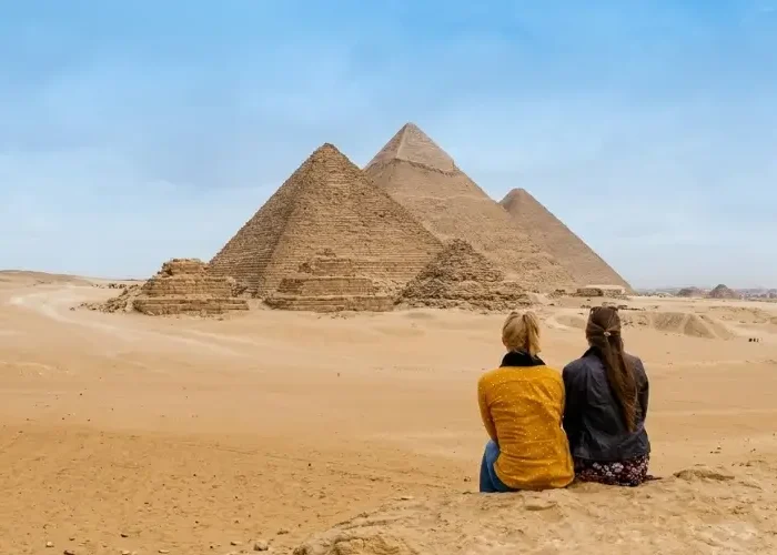 Two female travelers sitting on a hill and looking out at the Great Pyramids of Giza during a vacation planned for Egypt in 2026.
