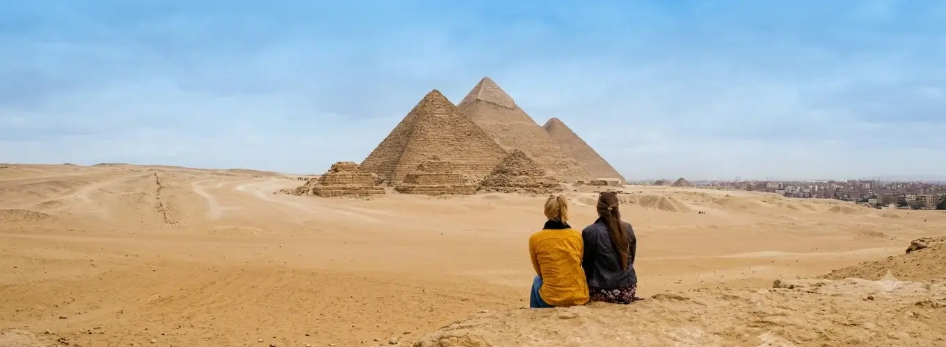 Two female travelers sitting on a hill and looking out at the Great Pyramids of Giza during a vacation planned for Egypt in 2026.