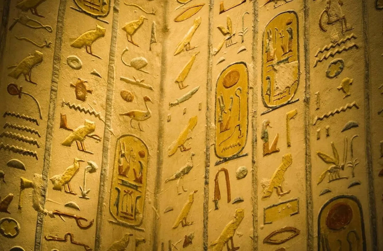 Detailed close-up of yellow and ochre colored Ancient Egypt symbols, featuring royal cartouches and hieroglyphs carved into a stone wall.