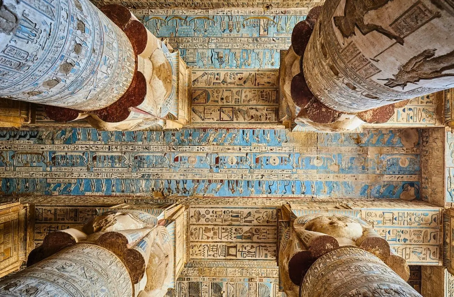 Looking up at the vibrant blue astronomical ceiling and massive stone columns carved with Ancient Egypt symbols and the face of the goddess Hathor at Dendera Temple.