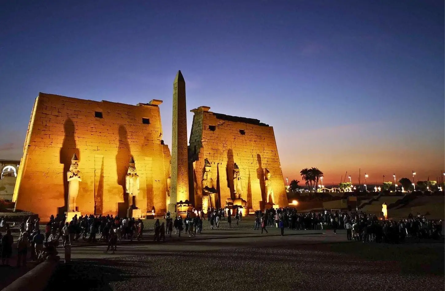 The illuminated entrance pylon and obelisk of the Luxor Temple in Egypt at twilight, with a crowd of tourists gathered in front.