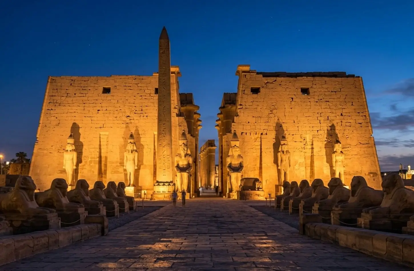 illuminated-luxor-temple-pylon-statues Luxor Temple night tour view showing the illuminated entrance pylon, ancient statues, and the Avenue of Sphinxes under a blue twilight sky.