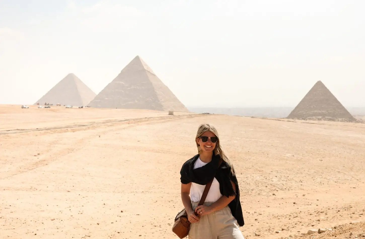 A smiling female solo traveler stands in the desert plateau with the iconic Pyramids of Giza rising majestically behind her during a trip to Egypt in October