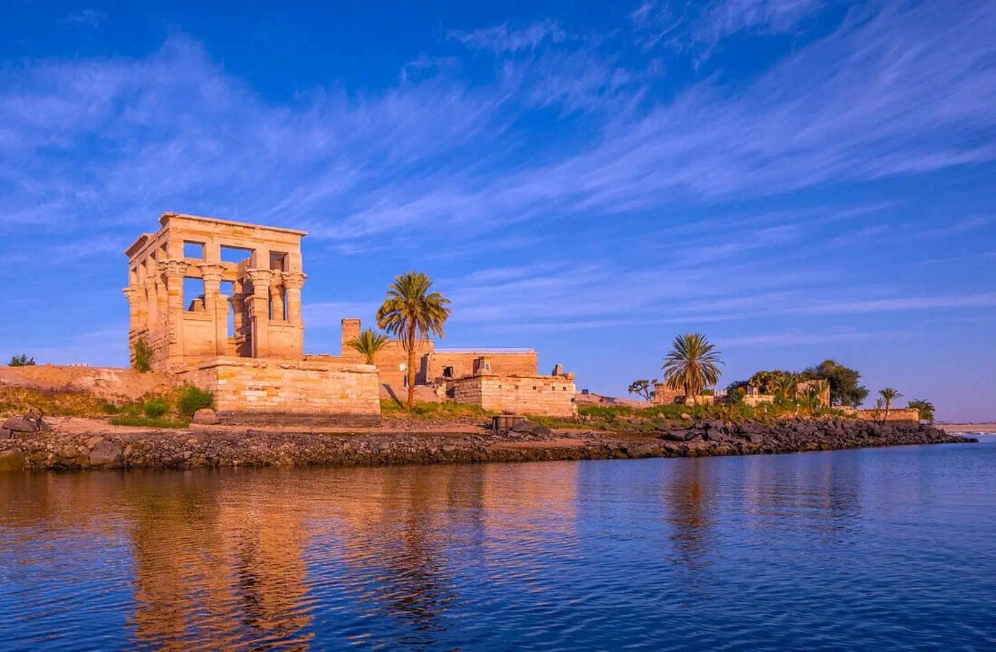 Trajan's Kiosk at the Philae Temple complex, situated on the edge of the Nile River with palm trees and a vibrant blue sky reflected in the water.