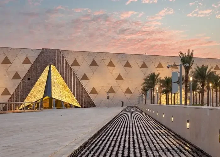A wide shot of the grand plaza and geometric stone architecture of the Grand Egyptian Museum complex during a vibrant dusk sunset in Egypt.