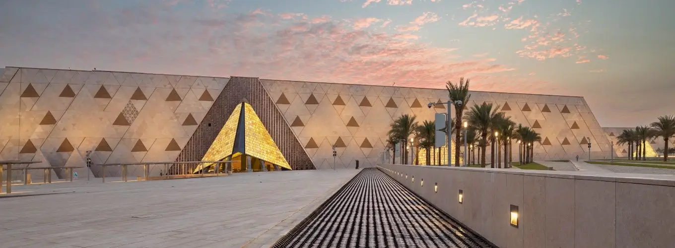 beautiful-sunset-at-grand-egyptian-museum-exterior A wide shot of the grand plaza and geometric stone architecture of the Grand Egyptian Museum complex during a vibrant dusk sunset in Egypt.