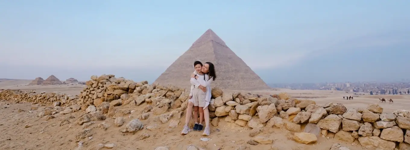 egypt-itinerary-10-days-giza-pyramids-family A mother hugging her son while sitting on a stone wall in the desert with the majestic Great Pyramid of Giza behind them, a highlight of a family friendly Egypt itinerary 10 days