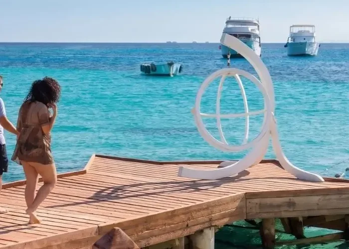 A couple enjoying a sunny vacation in Egypt in October, walking along a wooden pier over crystal-clear turquoise waters with boats in the background