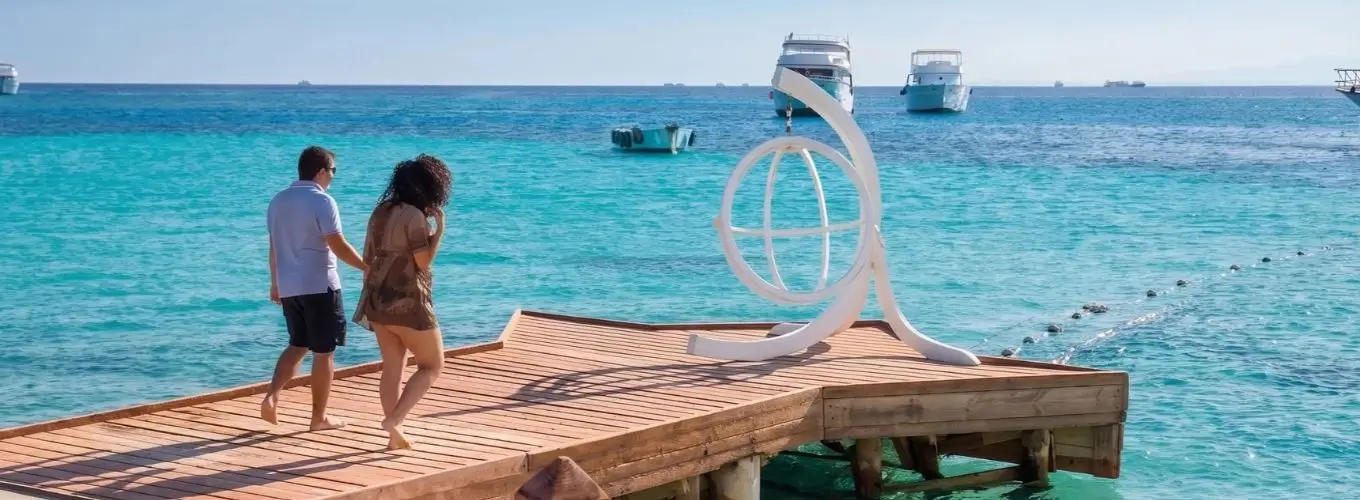 romantic-couple-walking-wooden-pier-resort A couple enjoying a sunny vacation in Egypt in October, walking along a wooden pier over crystal-clear turquoise waters with boats in the background