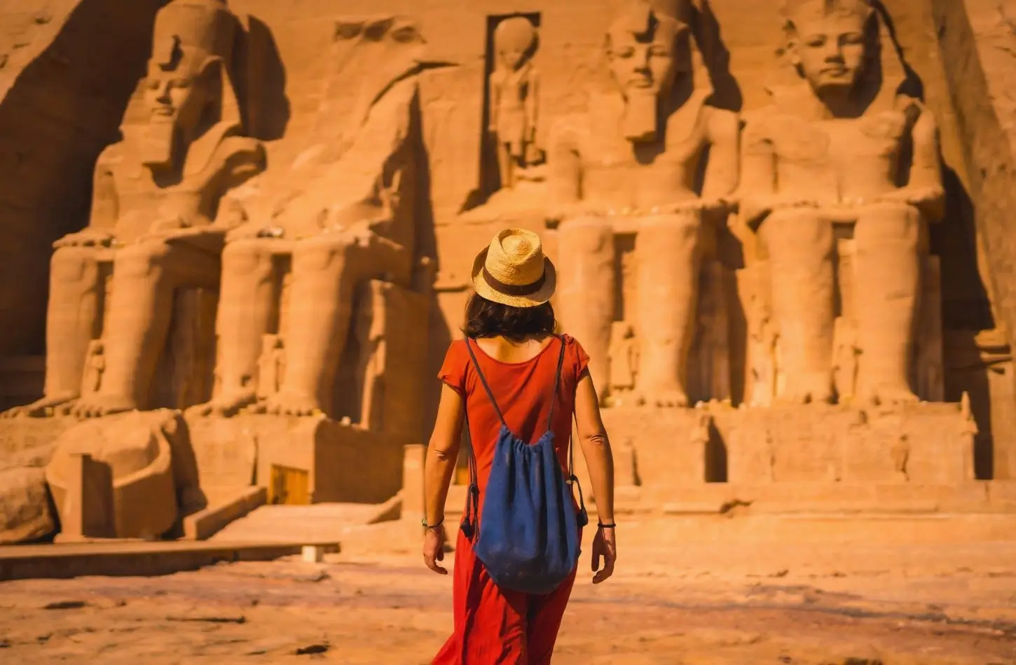 Egypt in September: A female traveler in a red dress, hat, and blue backpack looking up at towering ancient carved pharaoh statues.