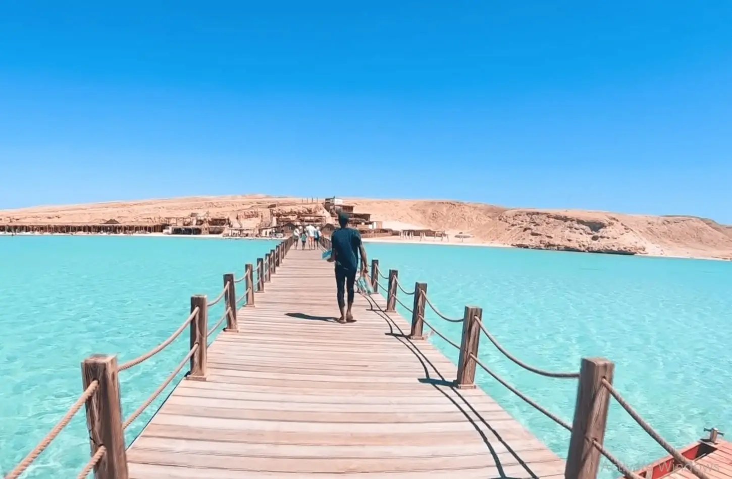 A traveler walking down a wooden pier over crystal-clear turquoise waters toward a sandy beach on Giftun Island, showcasing top island excursions and things to do in Hurghada.